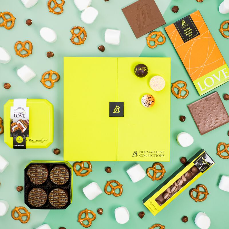 Overhead view of lime green signature truffle box with pieces of Hazelnut Bar, S'mores Love and Salted Caramel Pretzel Inclusion Bar. Ingredients such as retzels, marshmallows and hazelnuts surround chocolate box on a light green background. 